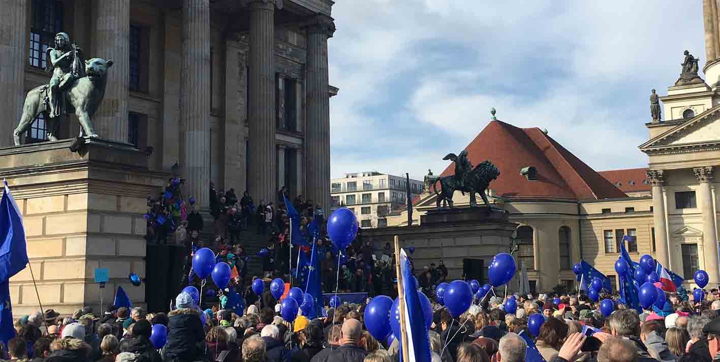 A crowd of people gathered outside a neo-classical building in a European city, holding royal blue balloons 