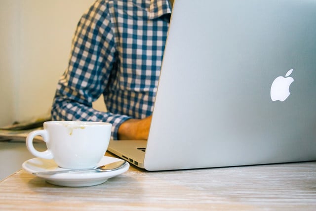 Man in a blue checked shirt sitting at a desk with a cup of coffee, working on his laptop