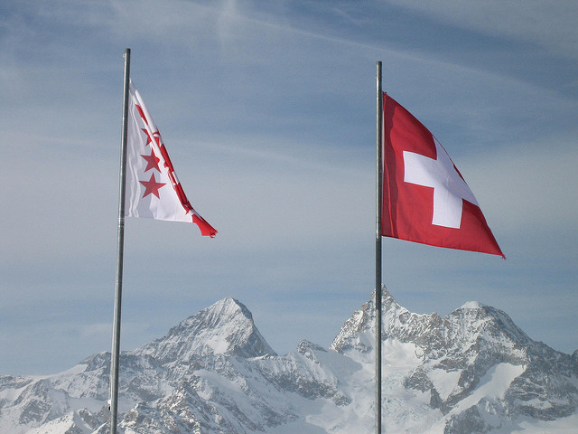 A raised Swiss flag seen in front of the snow-capped alps, waving gently in the breeze 